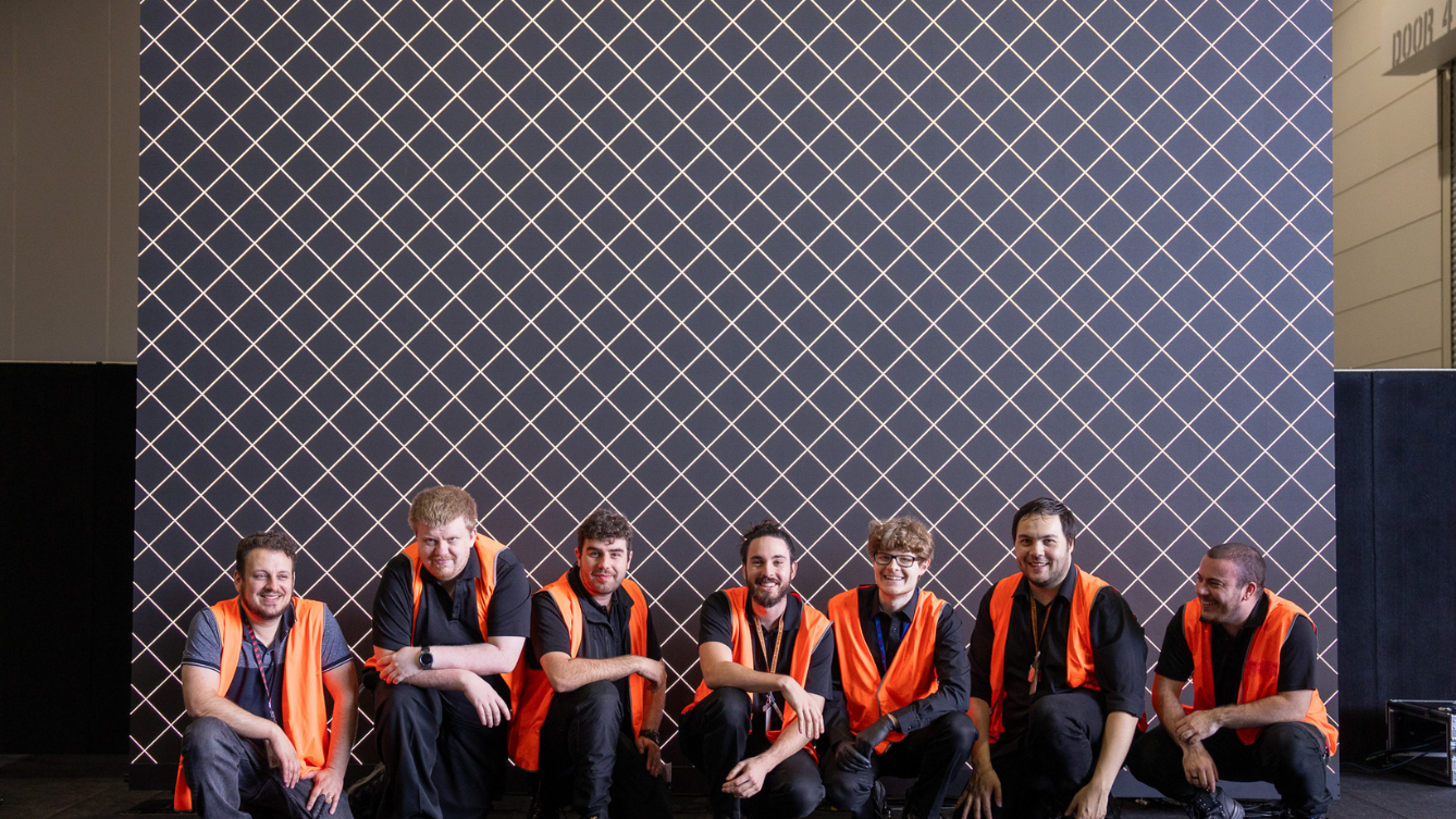 A row of people in high-vis orange vests. They are kneeling on the ground in a row in front of a large backdrop with a grid design. 