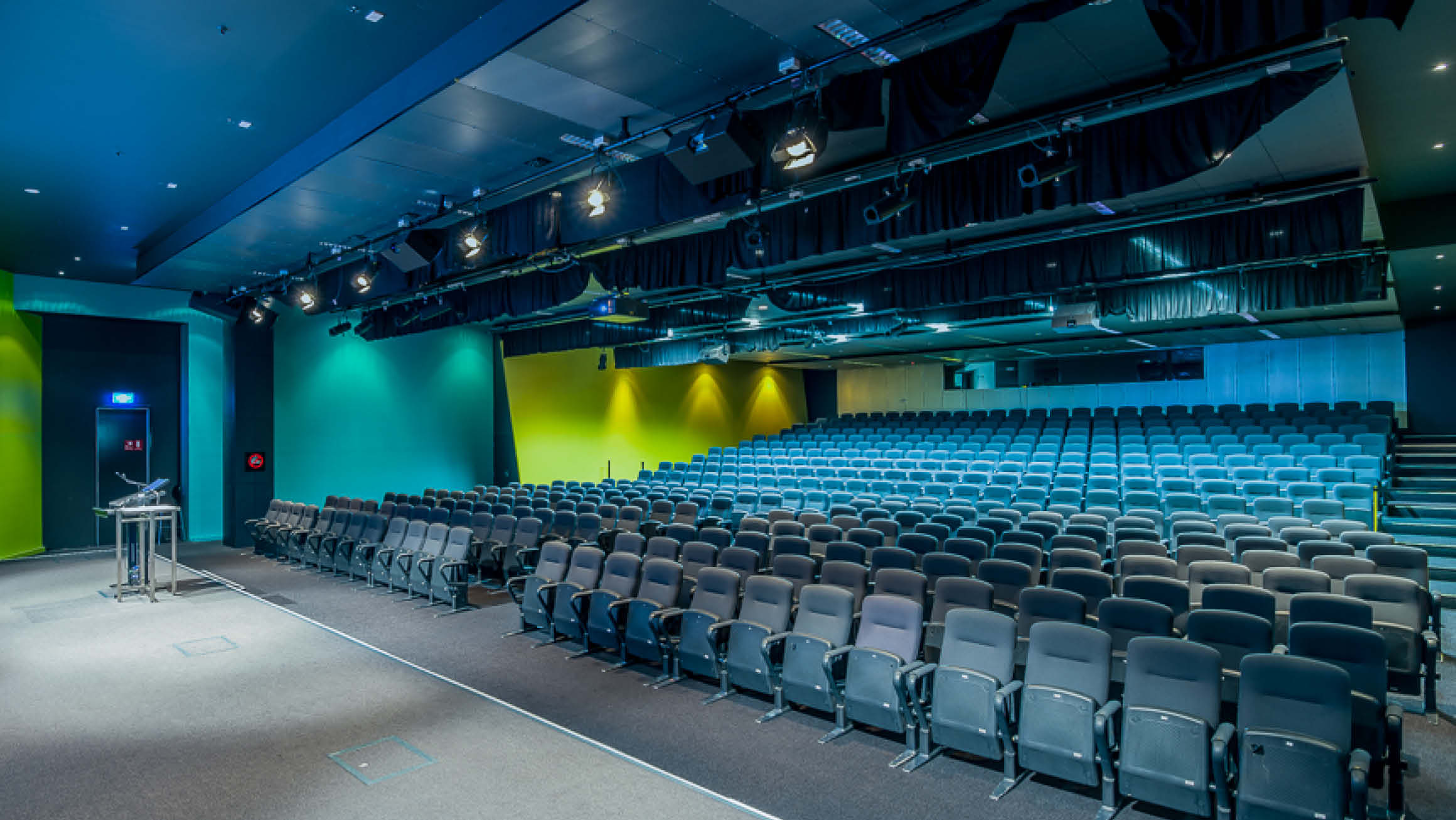 Large empty theatre with rows of chairs facing an empty stage with a lectern on it. The room is under blue and green lighting. 