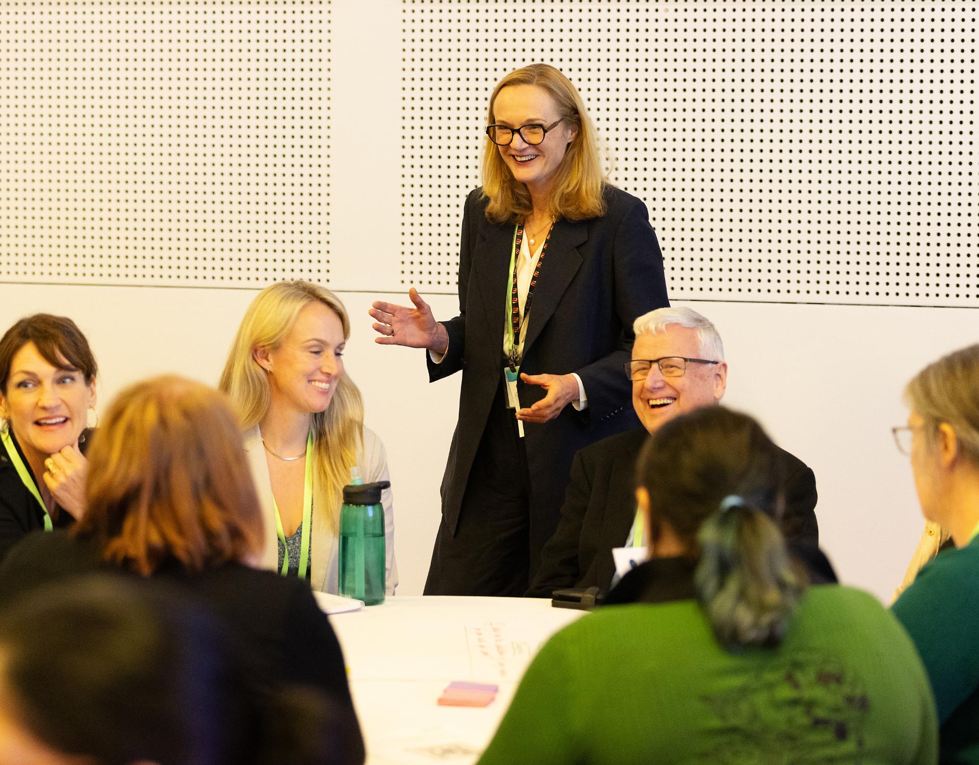 Woman is standing in black suit in front of a table of people. They are smiling and engaged in conversation. 