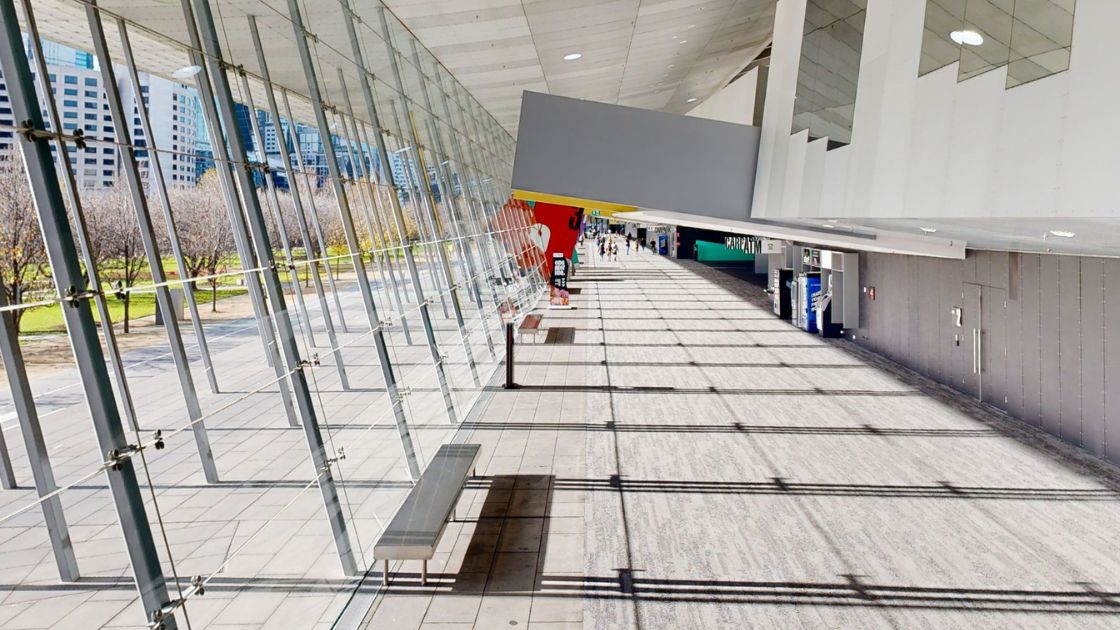 View of a large walkway with grey carpet. Large double story windows line one side with light streaming through. Metal poles that support the windows cast a grid layout of shadows onto the carpet. 