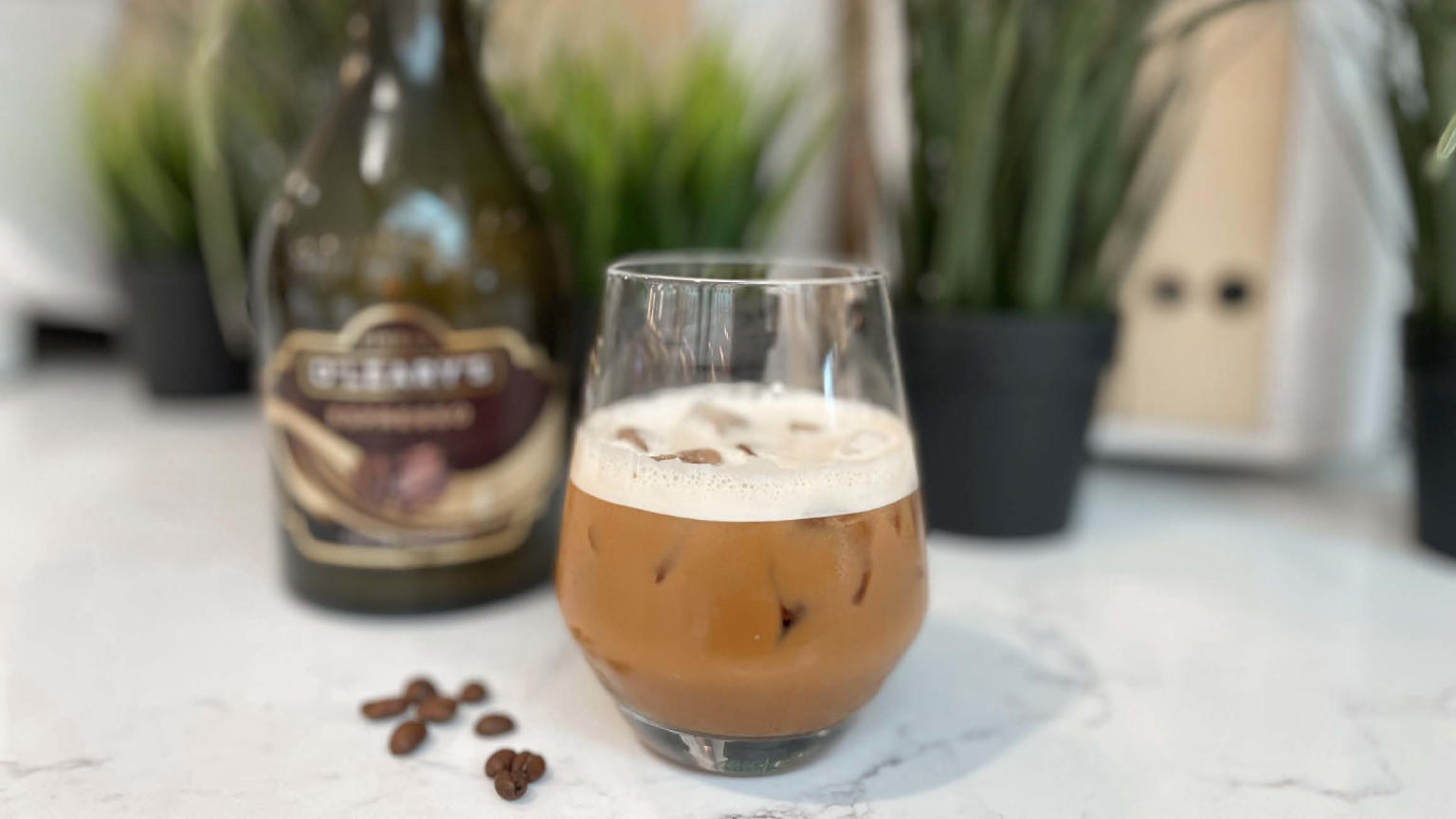 A delectable brown cocktail served in a short round glass, adorned with froth and accompanied by ice. Adjacent to it, a scattering of aromatic coffee beans adds an enticing touch. In the background, a dark bottle and black potted plants create a sophisticated ambience.