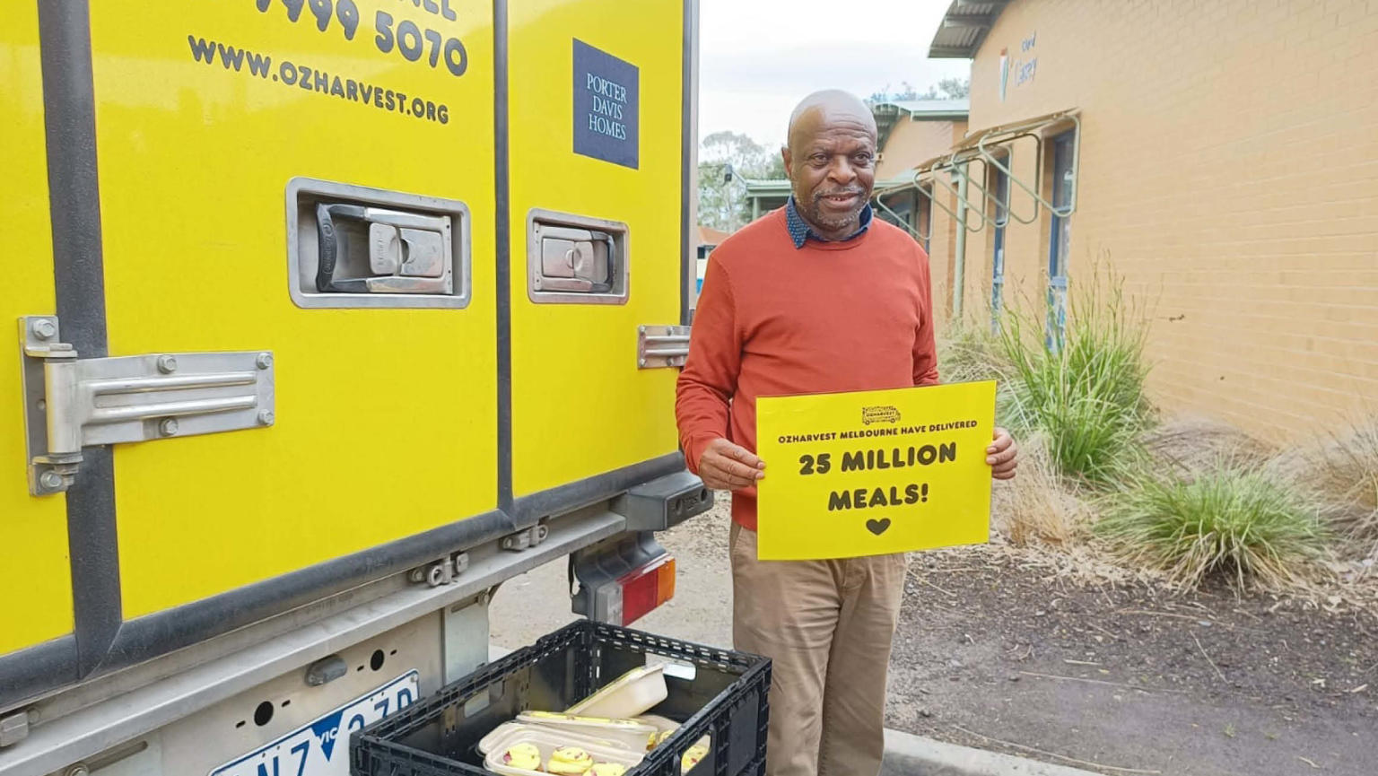 An image showing a man standing in front of a yellow OzHarvest van with a crate of food, holding a sign that says, 'OzHarvest Melbourne have delivered 25 million meals.' 