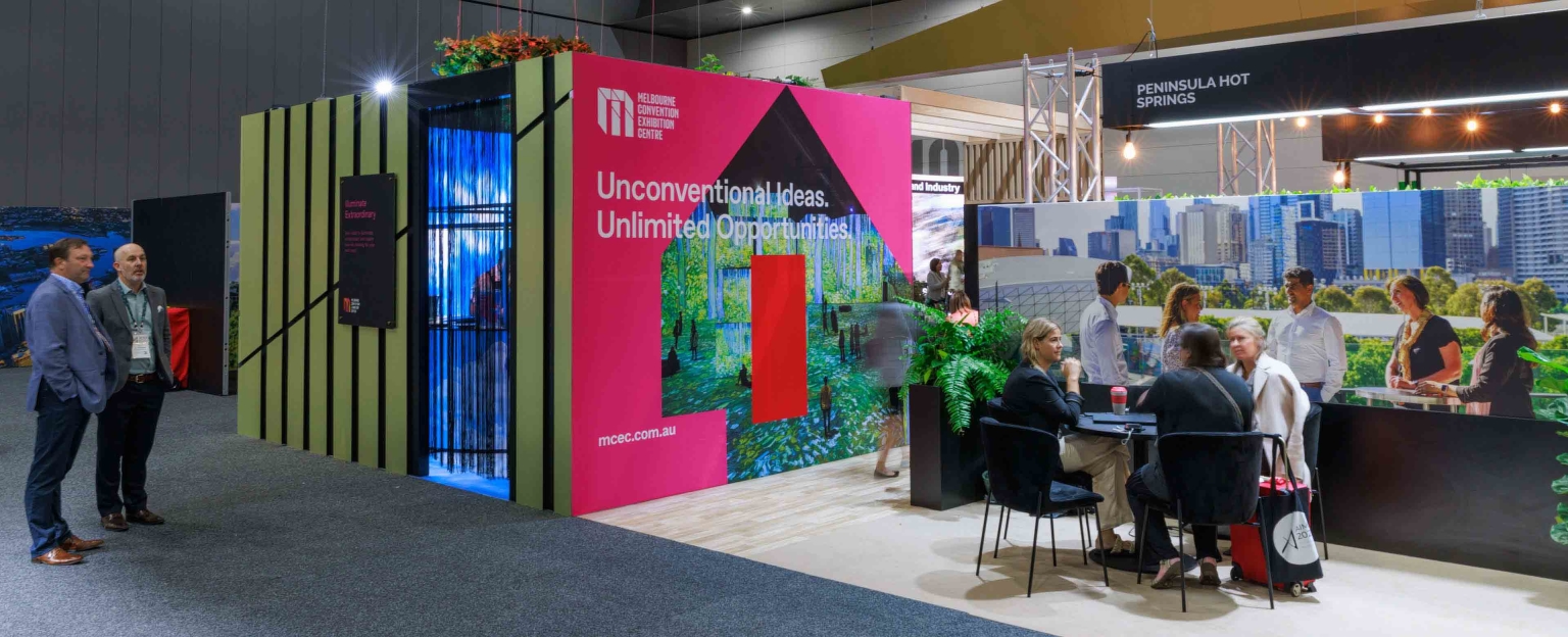 Melbourne Convention and Exhibition Centre exhibition stand at AIME. The stand is square, with one gold wall and an entryway, and one pink wall with a cut-out shape. There are people gathered around a table nearby, and others walking past.
