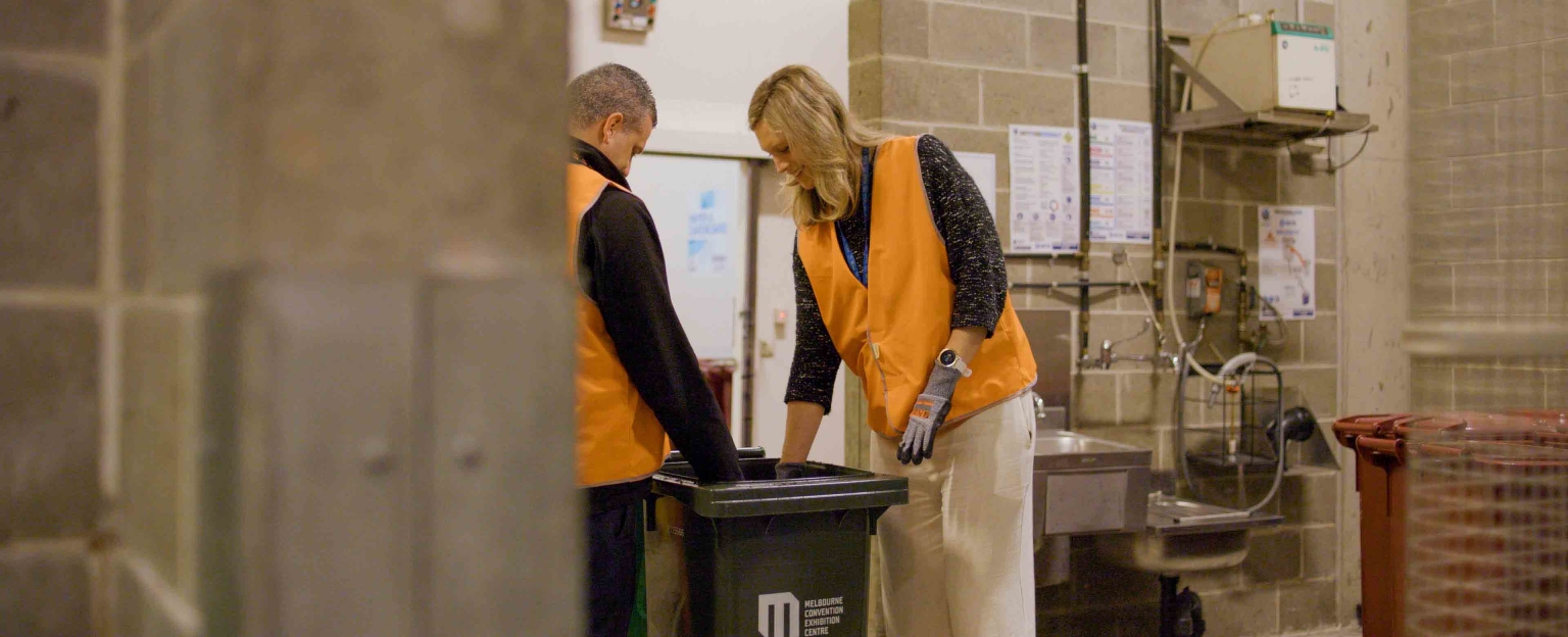 Sam Ferrier, Sustainability Manager at Melbourne Convention and Exhibition Centre talking with a man, both in orange high-visibility vests and gloves are standing in front of an organic dehydrator. They are looking into a bin of organic waste and appear to be discussing it.