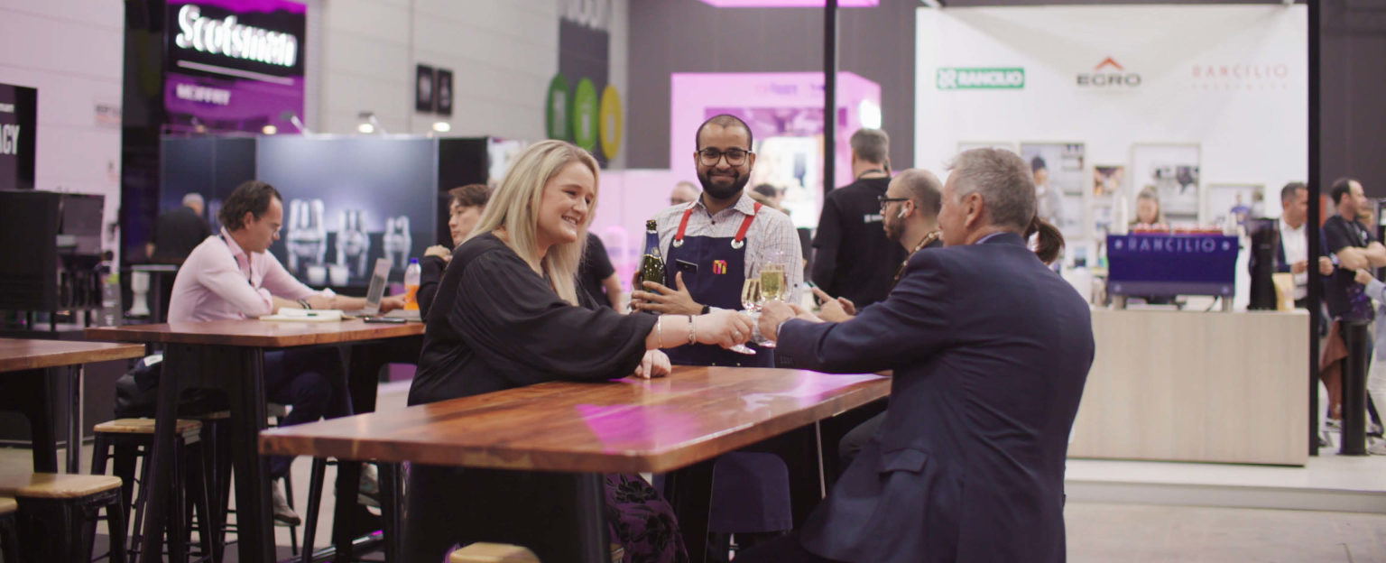 Woman sitting down at a table talking to a man in a suit with a waiter standing behind the table delivering drinks. A busy exhibition unfolds behind them with stands and people walking around. 