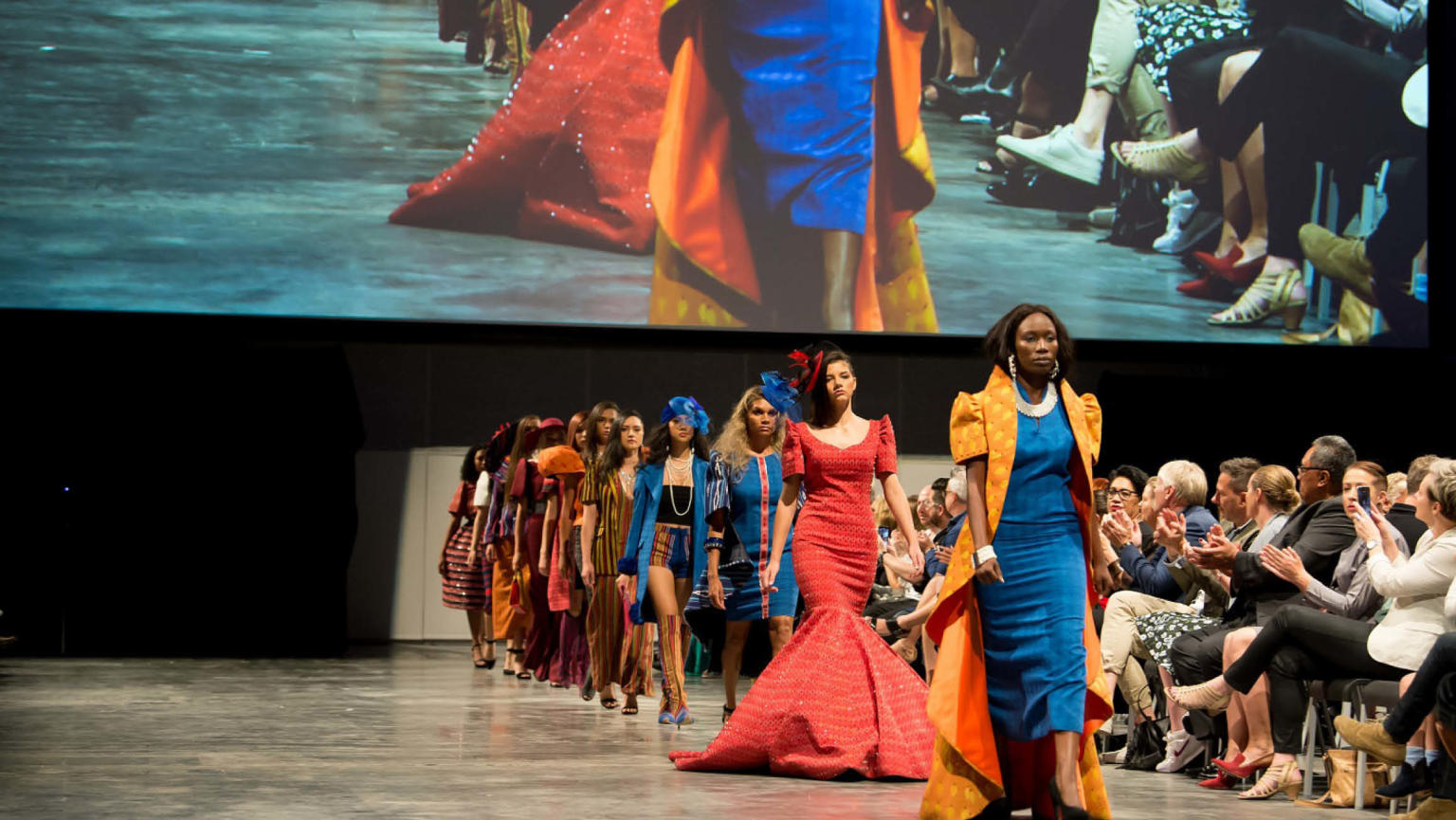 Models dressed in colourful clothing walking on a catwalk, while a large screen serves as the backdrop. The audience applauds in support.