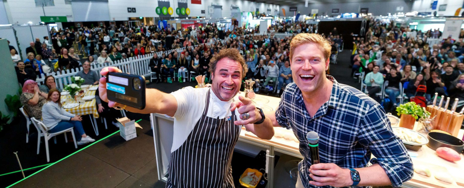 Chef Miguel Maestre and a man holding a microphone standing with their backs to a seated large crowd, taking a lively group selfie on a mobile phone.