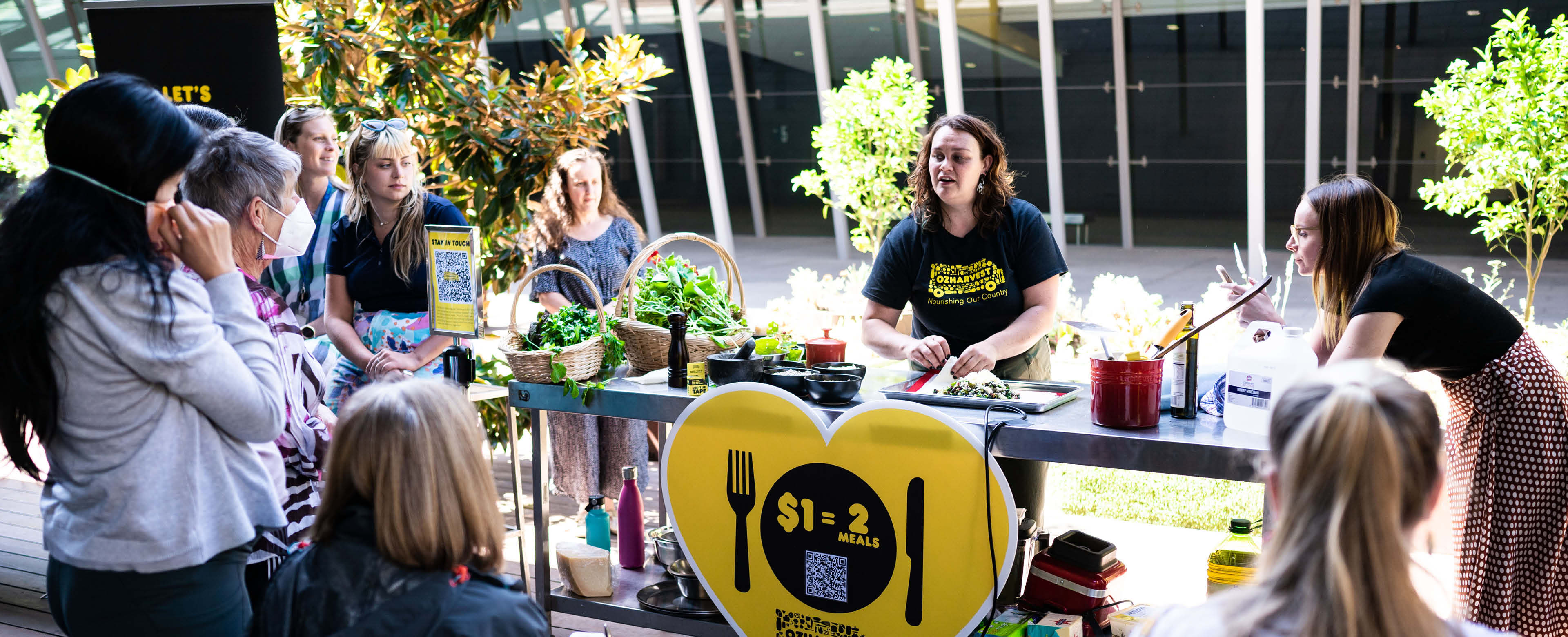 An image showing a woman demonstrating cooking a meal at a table outdoors surrounded by produce, with a group of people standing around the table and watching with curiosity. 