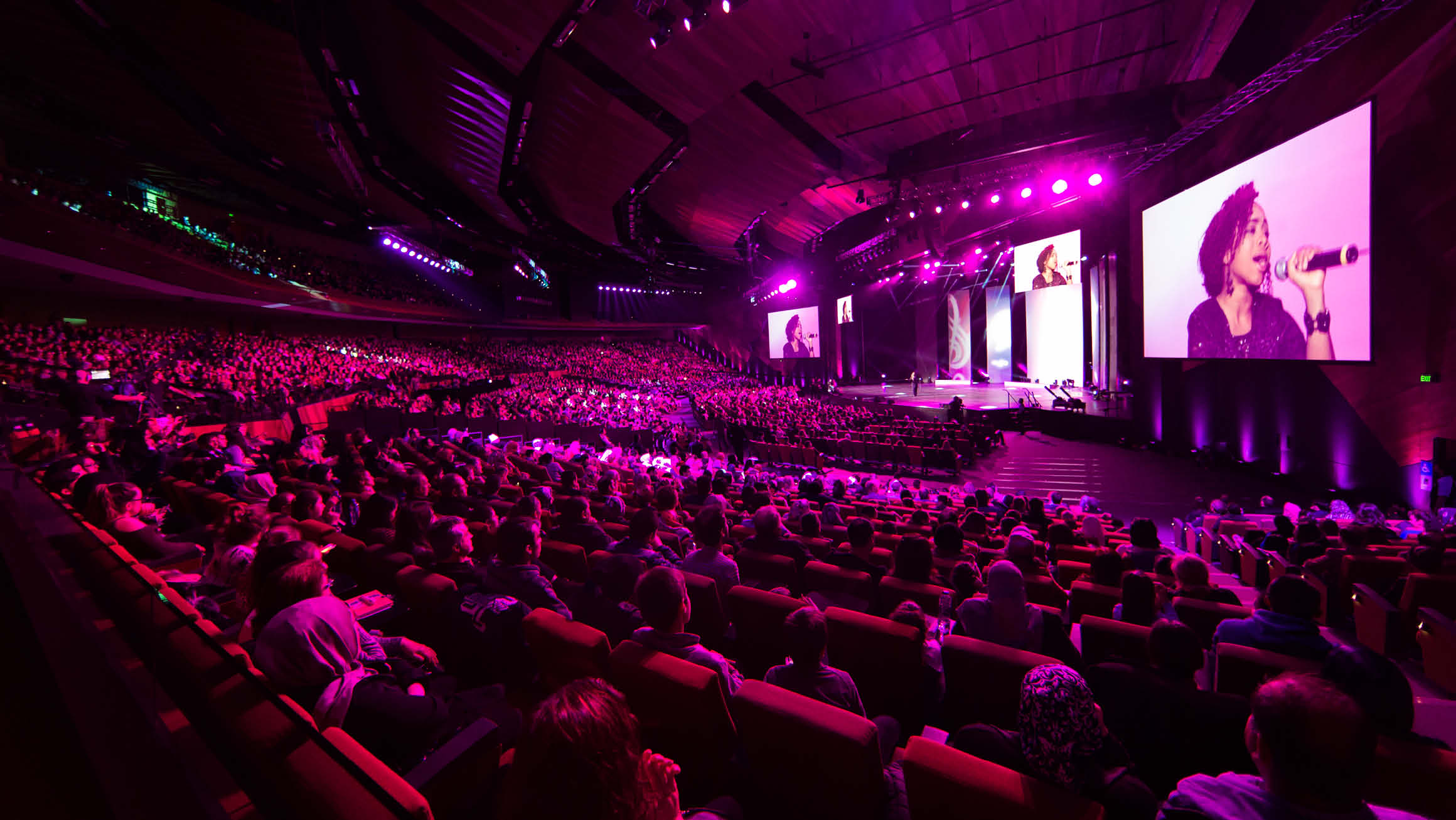 A large auditorium is filled with people watching a performance on stage. A woman is singing, and her image is projected onto two large digital screens to either side of the stage. The audience is captivated by the performance.