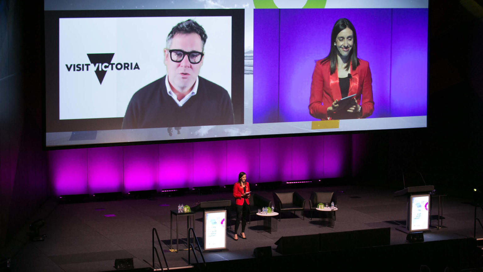 A woman confidently stands on a stage adorned with armchairs, tables, and a podium, dressed in a striking red blazer. Behind her, a sizeable digital screen displays a real-time feed of herself alongside a man speaking, accompanied by a logo next to him. The stage setup creates a dynamic and visually engaging backdrop for the speaker.