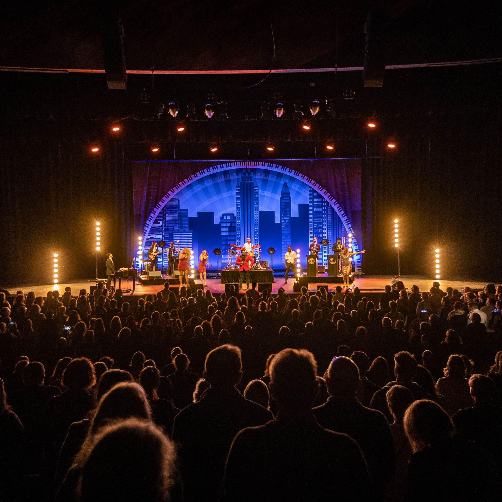 View from the back of a crowded room of people facing a lit up stage with a performance on. There is a blue semi circle backdrop behind the people on stage. 