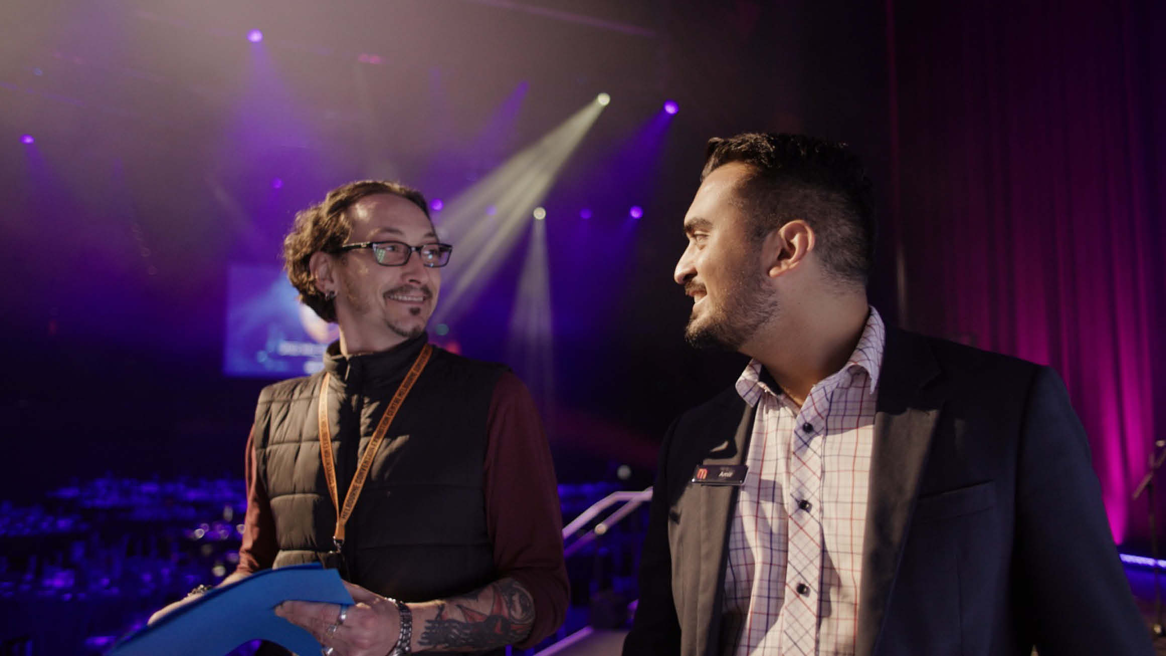 A man in a shirt and suit walks and engages in conversation at an event, accompanied by another individual wearing glasses, a puffer vest, and a shirt with a lanyard, holding a folder. The background features radiant lighting and floor-to-ceiling curtains, creating an elegant atmosphere.