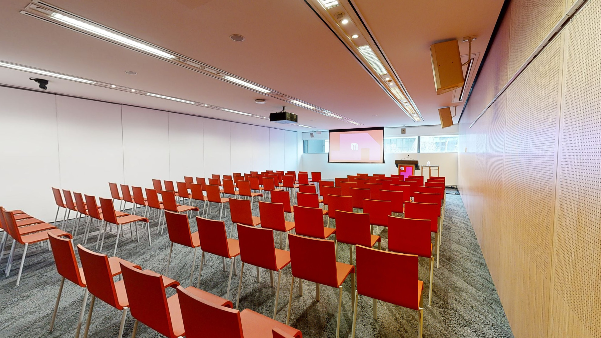 Conference or meeting room with orange chairs set up in rows facing a large projector screen with a strip of windows sitting behind at eye level. A lectern sits to the right of the screen. 