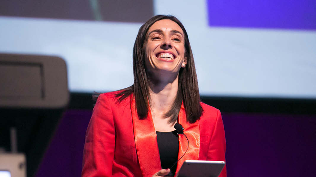 A confident woman wearing a vibrant red blazer stands at a podium, holding a tablet and addressing a crowd with a smile. The focus of her attention is behind the camera, creating an engaging and energetic atmosphere.