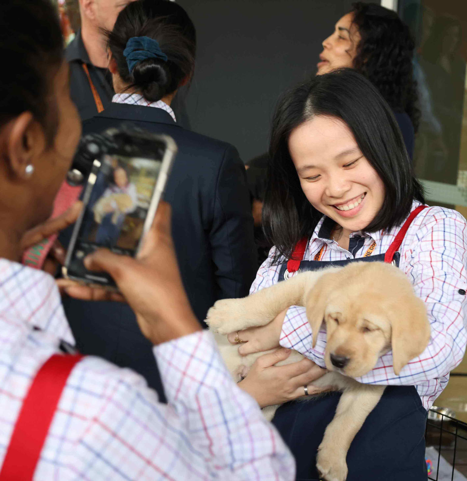 A woman holding a golden retriever puppy is smiling while looking at the puppy. Another woman in the foreground is seen taking a picture of them. 