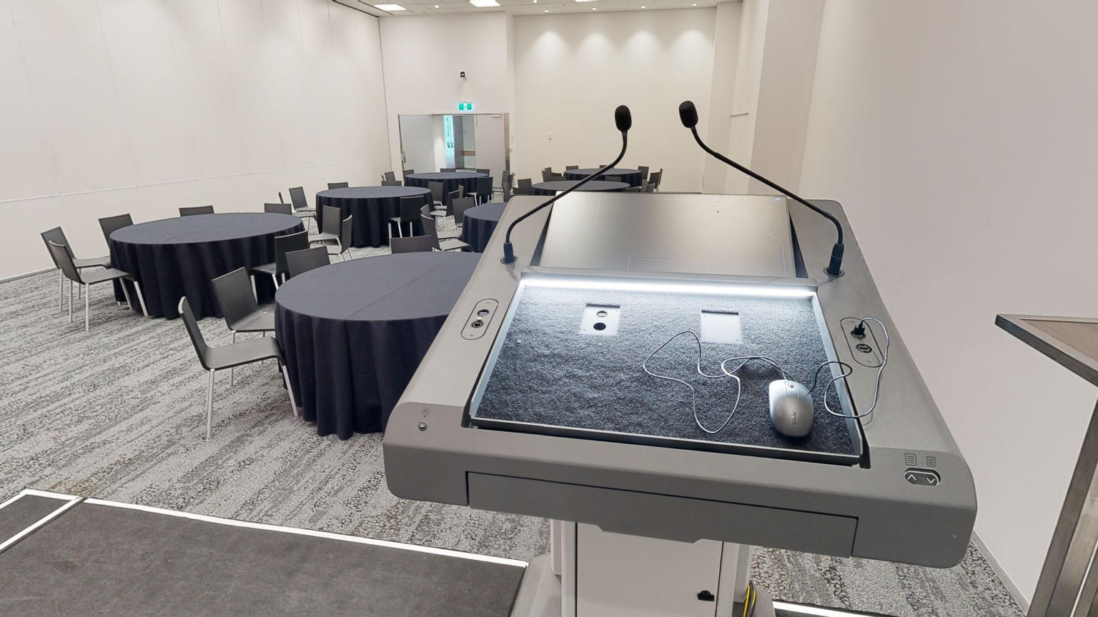 A professional conference or meeting room with neatly arranged round tables and chairs. A lectern and stage sit to the side of the room facing out towards the tables. 