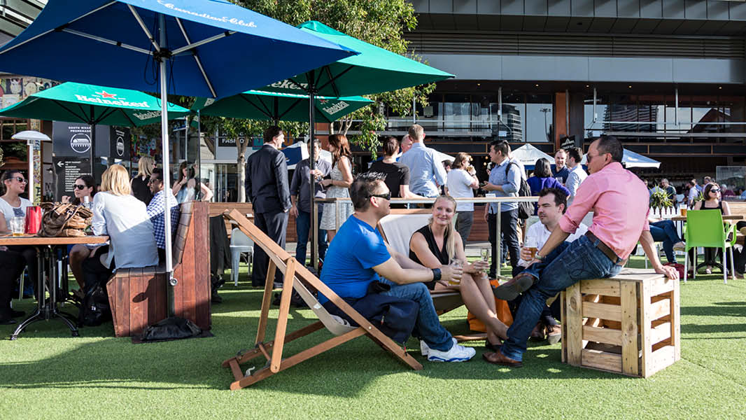 A lively scene showcasing multiple groups of people enjoying the outdoors, seated on a well-maintained lawn. Some are seated on wooden chairs, some are sitting at wooden tables, all neatly arranged under sun shade umbrellas shielding them from the sun. 