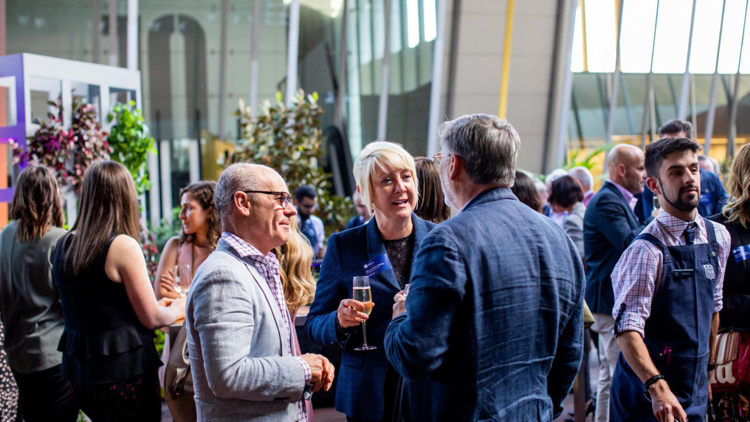 Three people stand in the foreground holding champagne talking to one another. A busy crowd is behind them and they are outside with the MCEC building slightly in view in the background. 