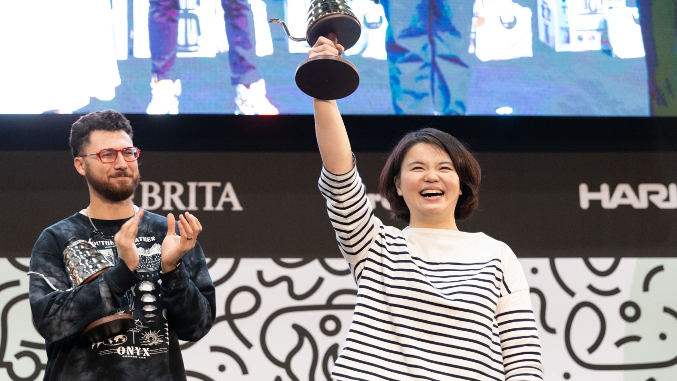 Man standing in the background applauding a women in the foreground holding up trophy. She is smiling. 