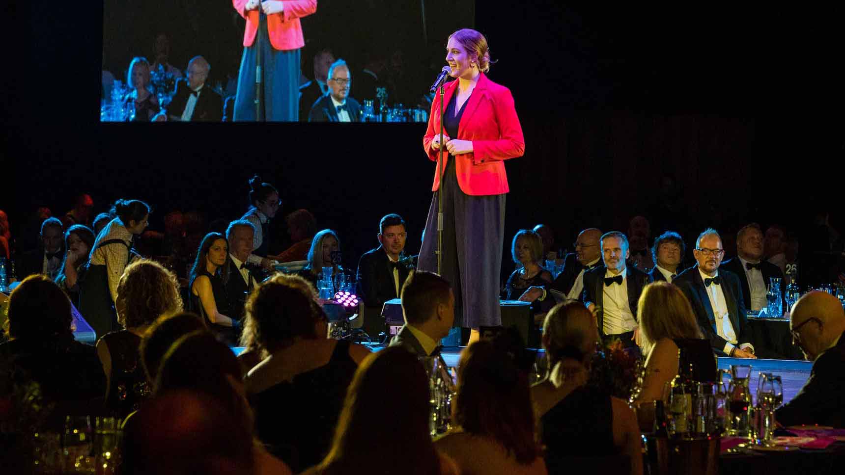 A woman standing on a stage in front of a crowd who are seated at tables in formal attire. 