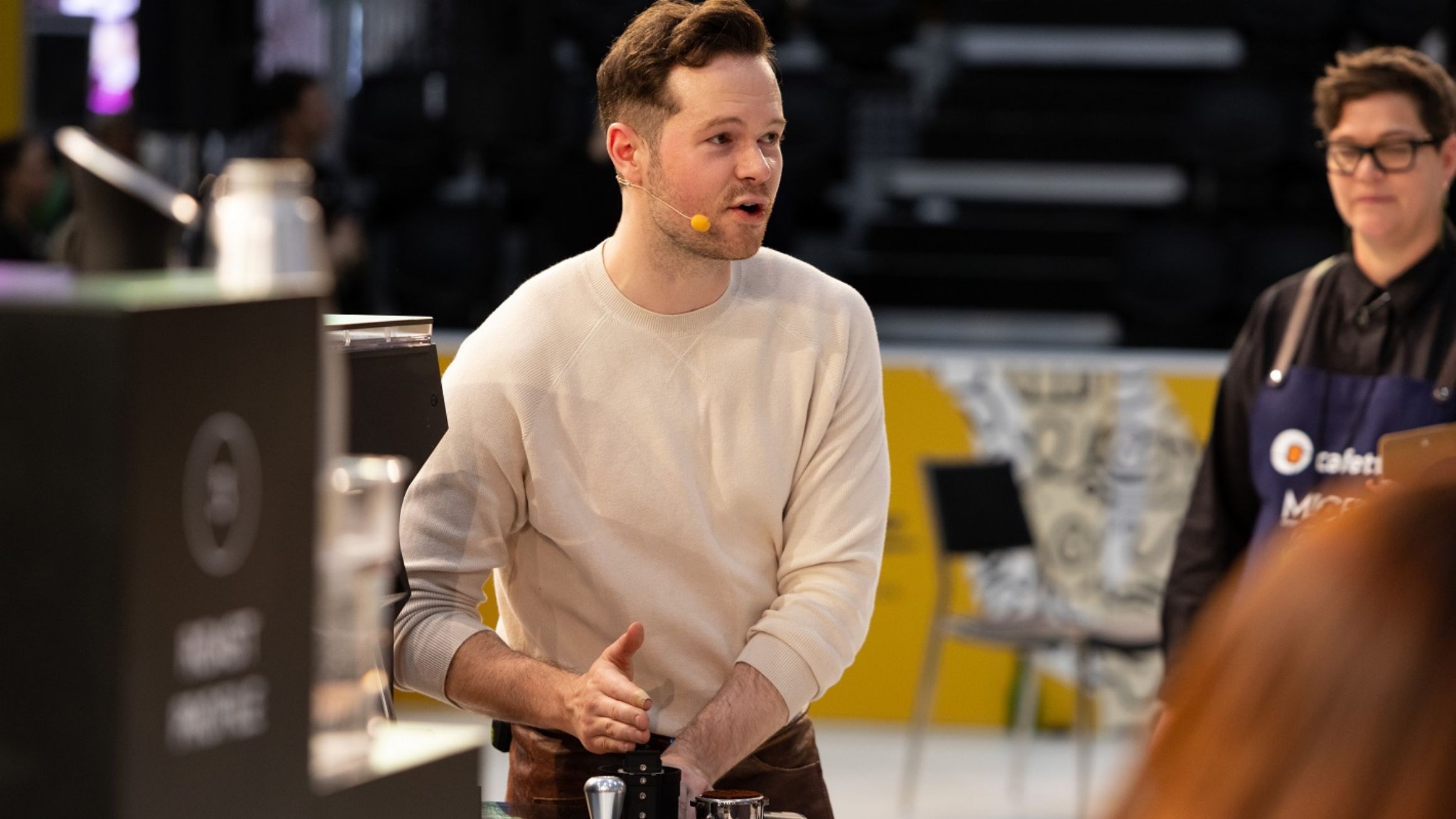 A man in a white jumper speaking into a microphone head piece facing a crowd off screen. He is running a demonstration. 