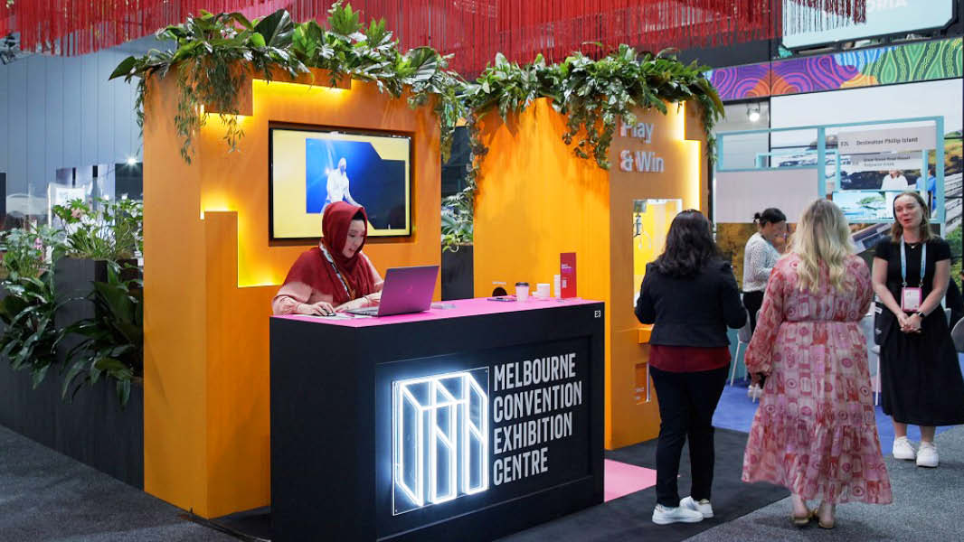 An eye-catching exhibition stand with a captivating arrangement: long red fringing suspended from the ceiling, an inviting orange wall featuring a walk-through cutout adorned with lush greenery, and a dynamic digital screen enhanced by neon lights.