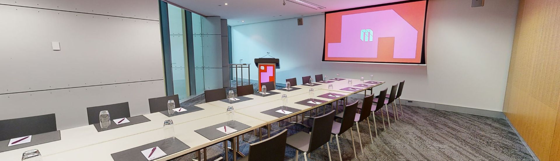 A conference or meeting room featuring a spacious boardroom layout equipped with a sizable screen and comfortable chairs for attendees. A lectern also sits in the corner to the left of the screen next to a large floor to ceiling window. 