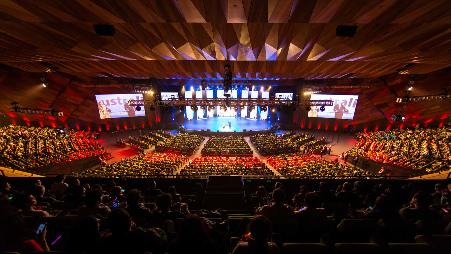 Image of a large multipurpose space within the Melbourne Convention and Exhibition Centre (MCEC). The space has ample multi-levelled green seating that faces a stage with two large digital screens, one on each side of the stage. The space is well-lit and an event is in progress.