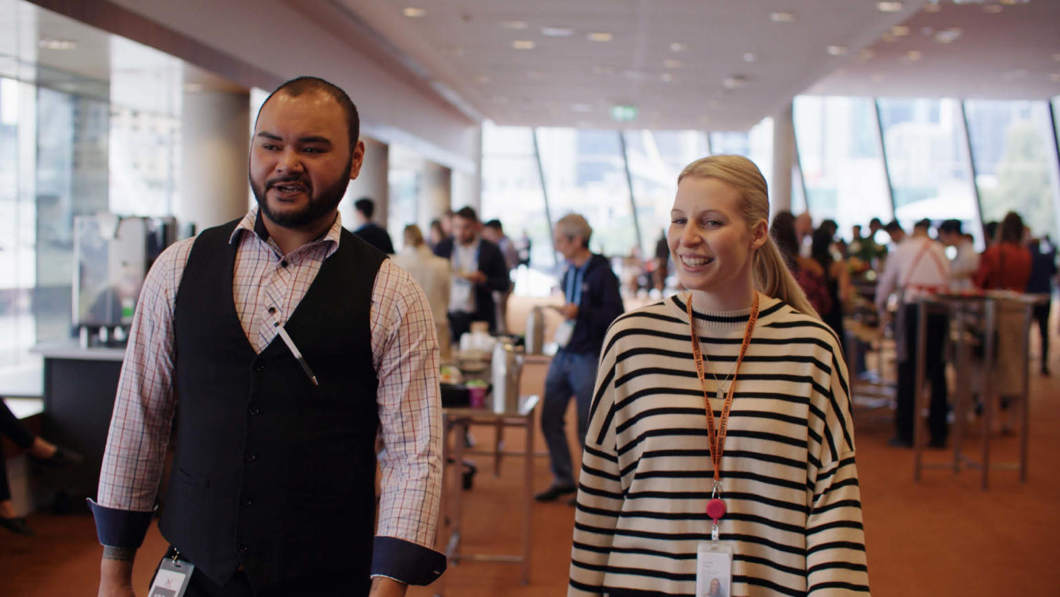 Anthea Fahey, wearing a striped jumper, walking alongside a man in a vest and shirt, moving away from a bustling event scene.