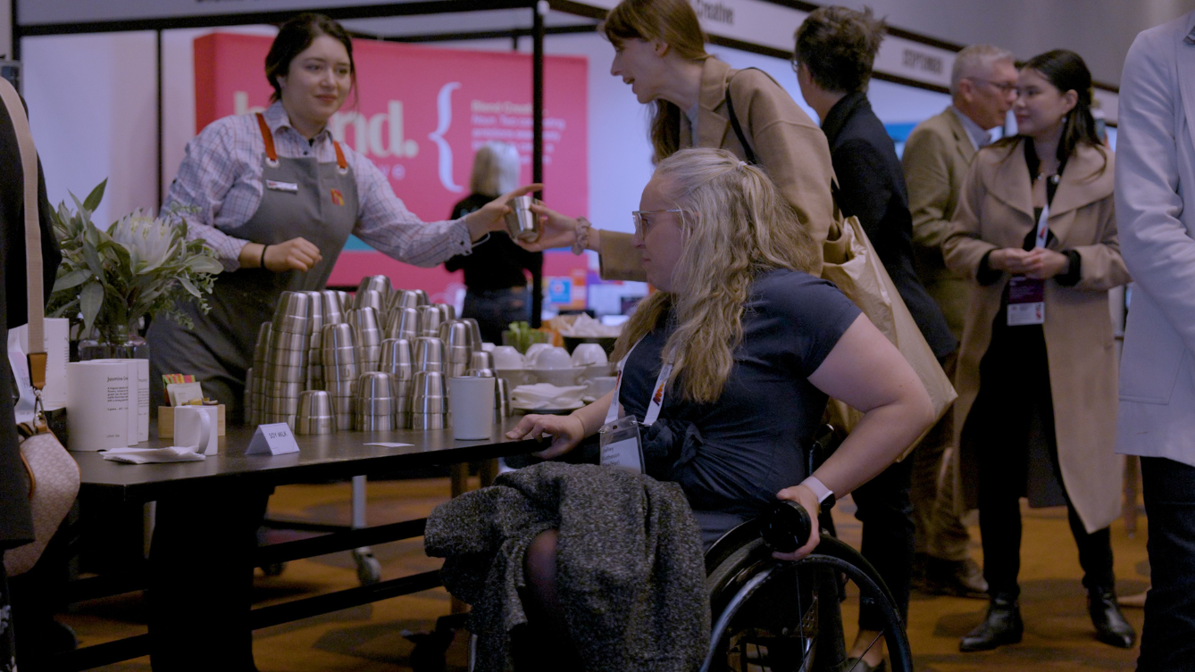 A women in a wheelchair is at the coffee stand. Another woman is standing behind the counter handing a cup to another person. 