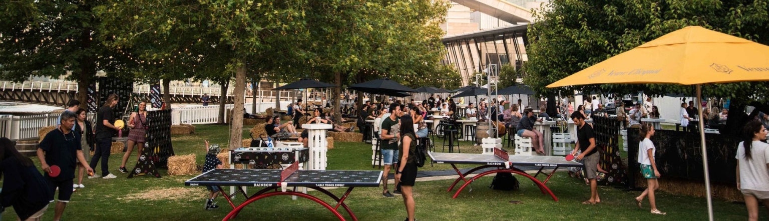 Outside grasses area with table tennis, umbrellas and tables and chairs set up with a crowd of people scattered throughout. Large buildings can be seen between the large green trees. 