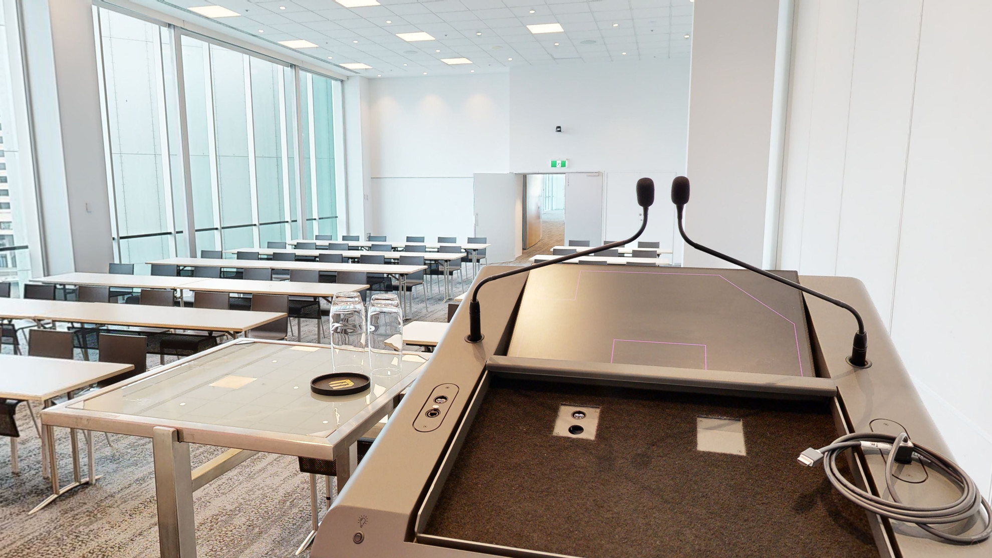 A professional conference or meeting room featuring a spacious layout, equipped with a stage and lectern and comfortable chairs for meetings and presentations. Table and chairs are set out in a classroom formation of rows. A large floor to ceiling window fills one wall of the room, which looks out to the Melbourne city. 