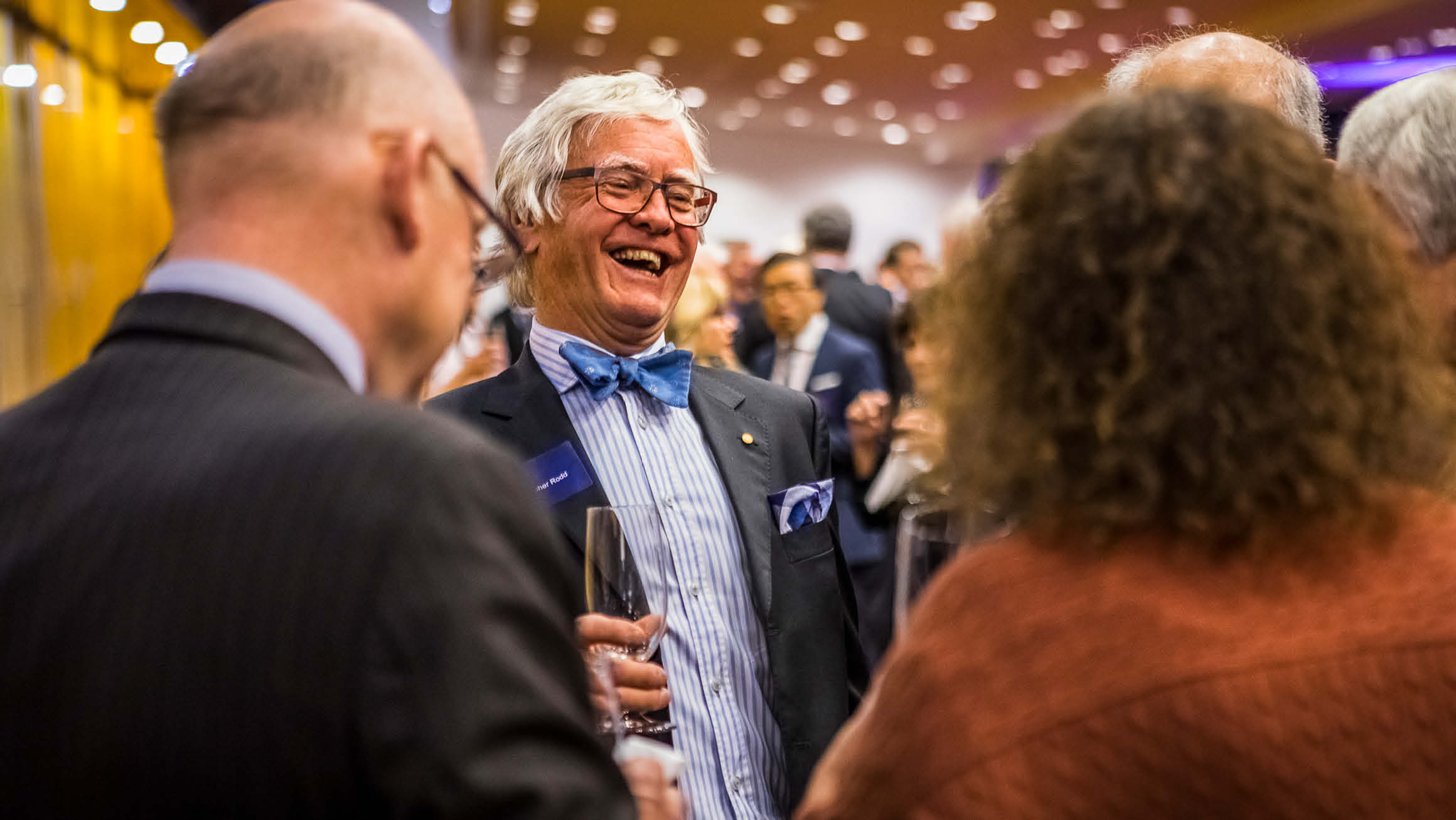 Man in suit and bow tie, stands in focus, laughing to two people in front of him. They have glasses in hand. 