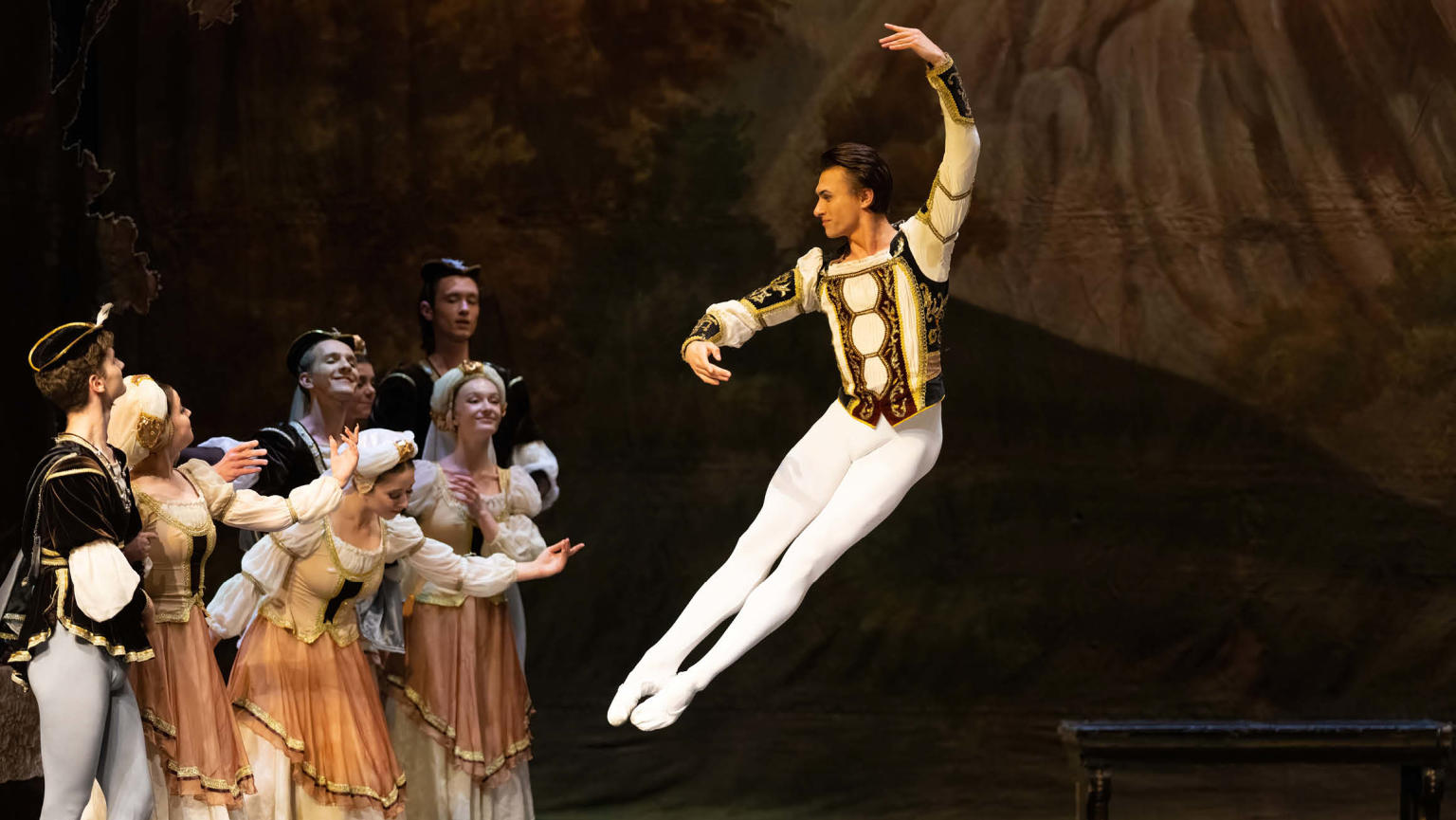 An image showing a group of ballerinas on a stage, gracefully dancing and attentively watching as a male danseur executes a jump, soaring gracefully into the air. 