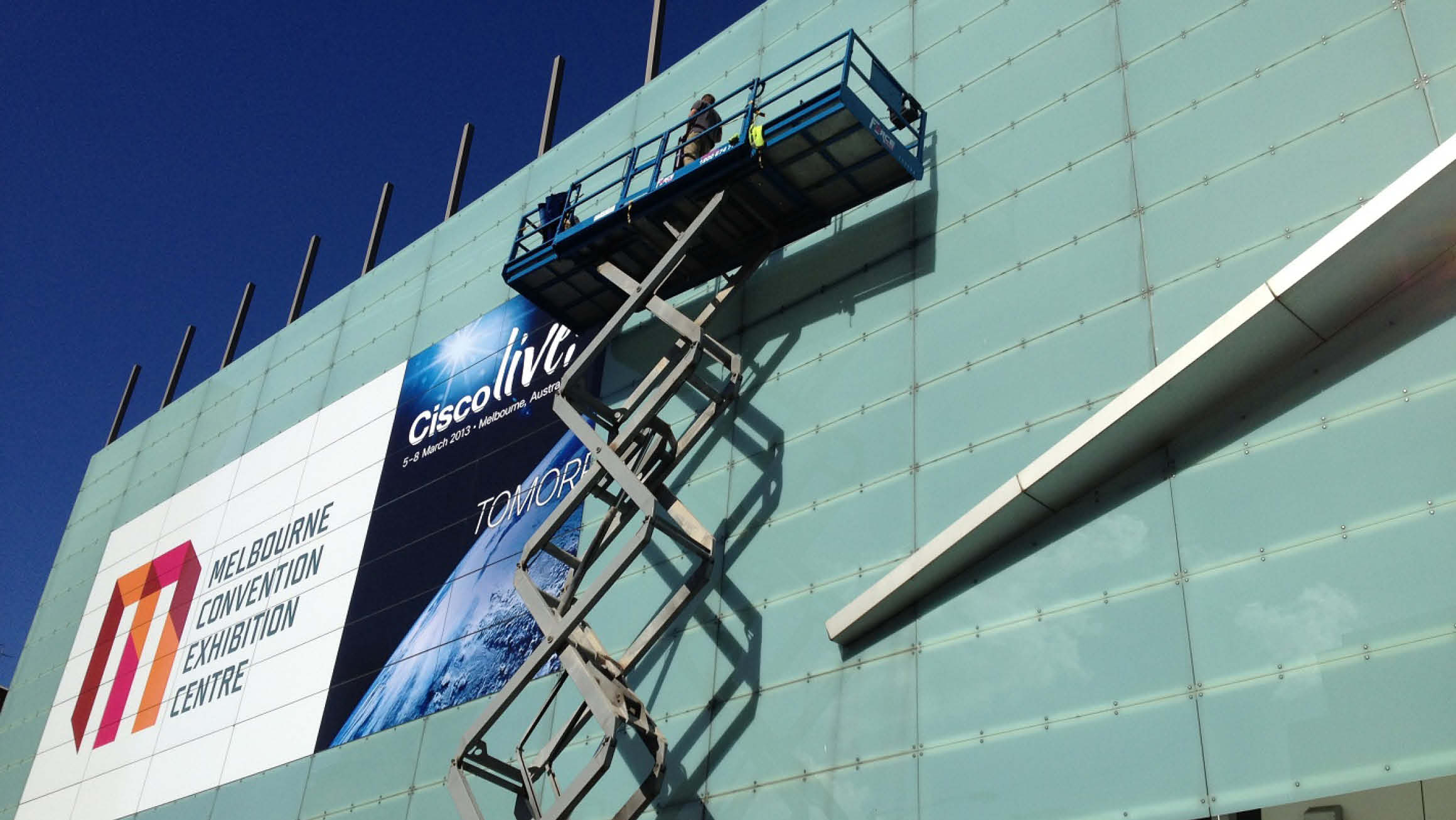 A building featuring a sizeable glass pane in the front, adorned with display stickers showcasing the text "Melbourne Convention Exhibition Centre." Adjacent to the existing stickers, an elevator scissor lift can be seen applying a new sticker next to the previous one.