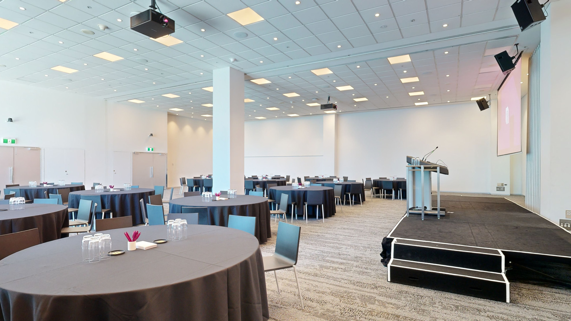 A spacious conference room with tables and chairs arranged for a meeting facing two large screens at the front of the room. Large floor to ceiling windows are to one side of the room. 