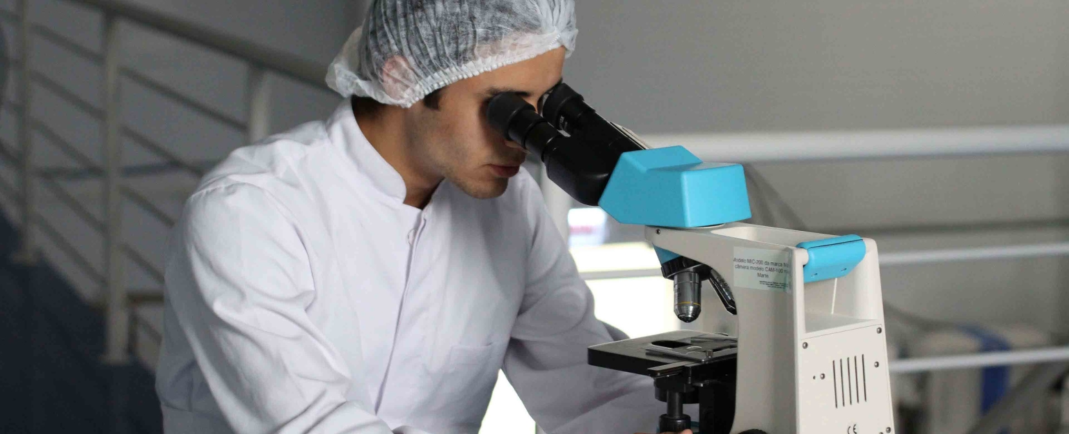 A scientist in a white lab coat and hair net is looking through a microscope. The background is a laboratory setting.
