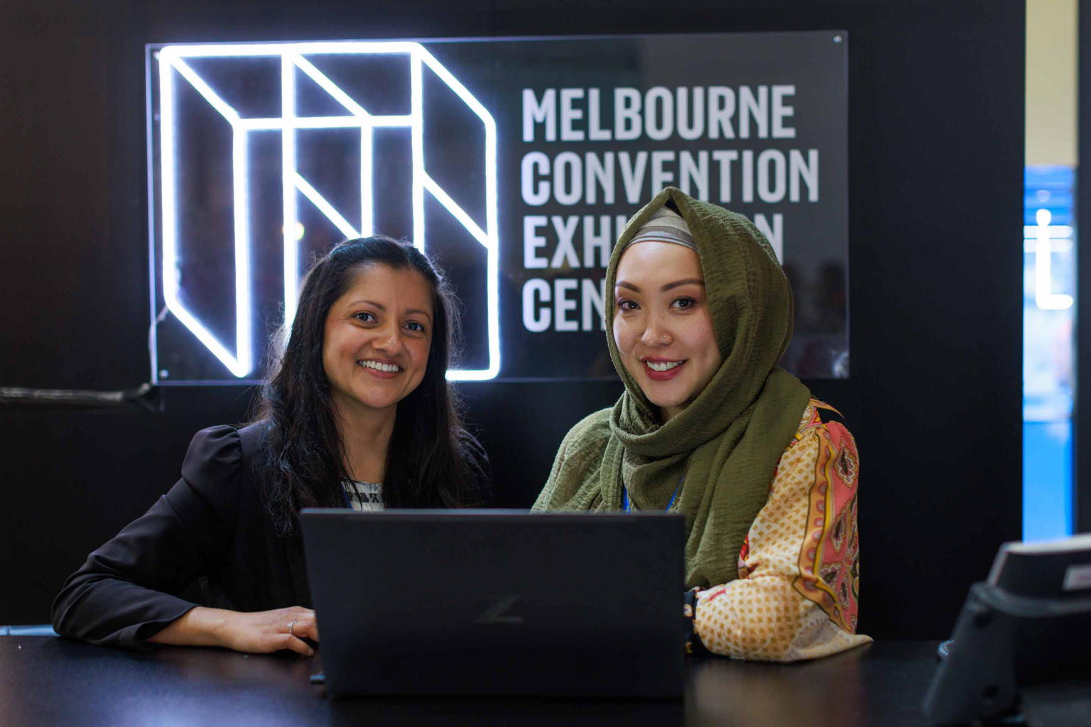 Two woman sitting at a table with a laptop in front of them. Behind them is a neon white sign of the Melbourne Convention and Exhibition logo on a black wall. 