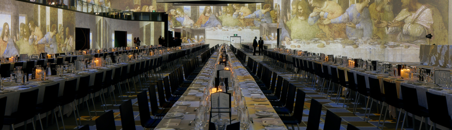 Large long tables running down a room. Perspective is from the head of the tables all beautifully set up with tableware and candles ready for a dinner. 