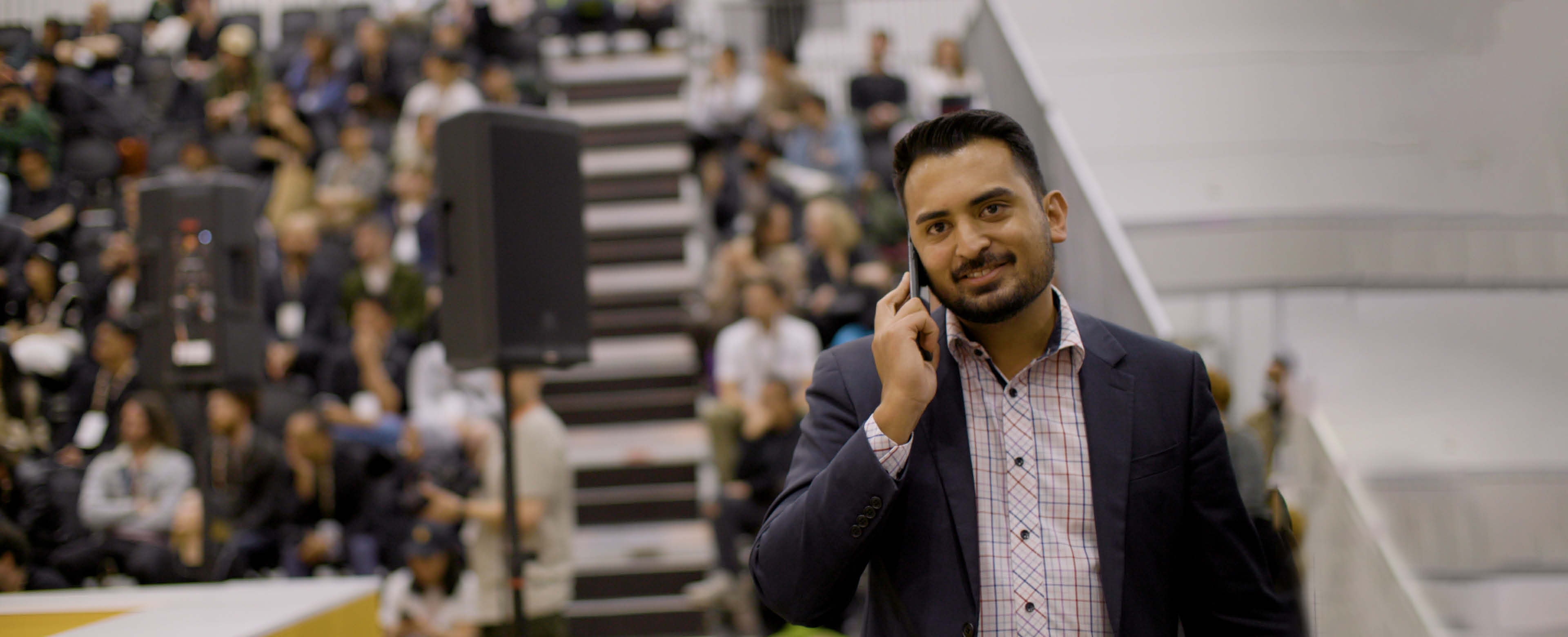 A man in a shirt and blazer happily talks on the phone while looking towards the camera. In the background, a sizeable crowd is seated on levelled stands, facing large speakers positioned before them.