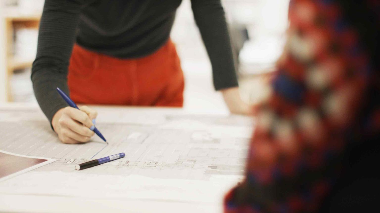 A person in red pants seen only from the shoulders down is leaning over a table with a pen in hand, drawing on large floor plan. 