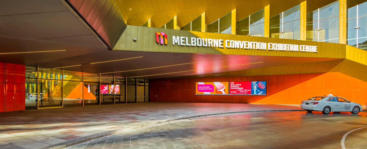 A large gold building featuring digital signage on the entrance wall, showcasing vibrant displays. A prominent logo is positioned on a protruding block, proudly displaying the words "Melbourne Convention Exhibition Centre."