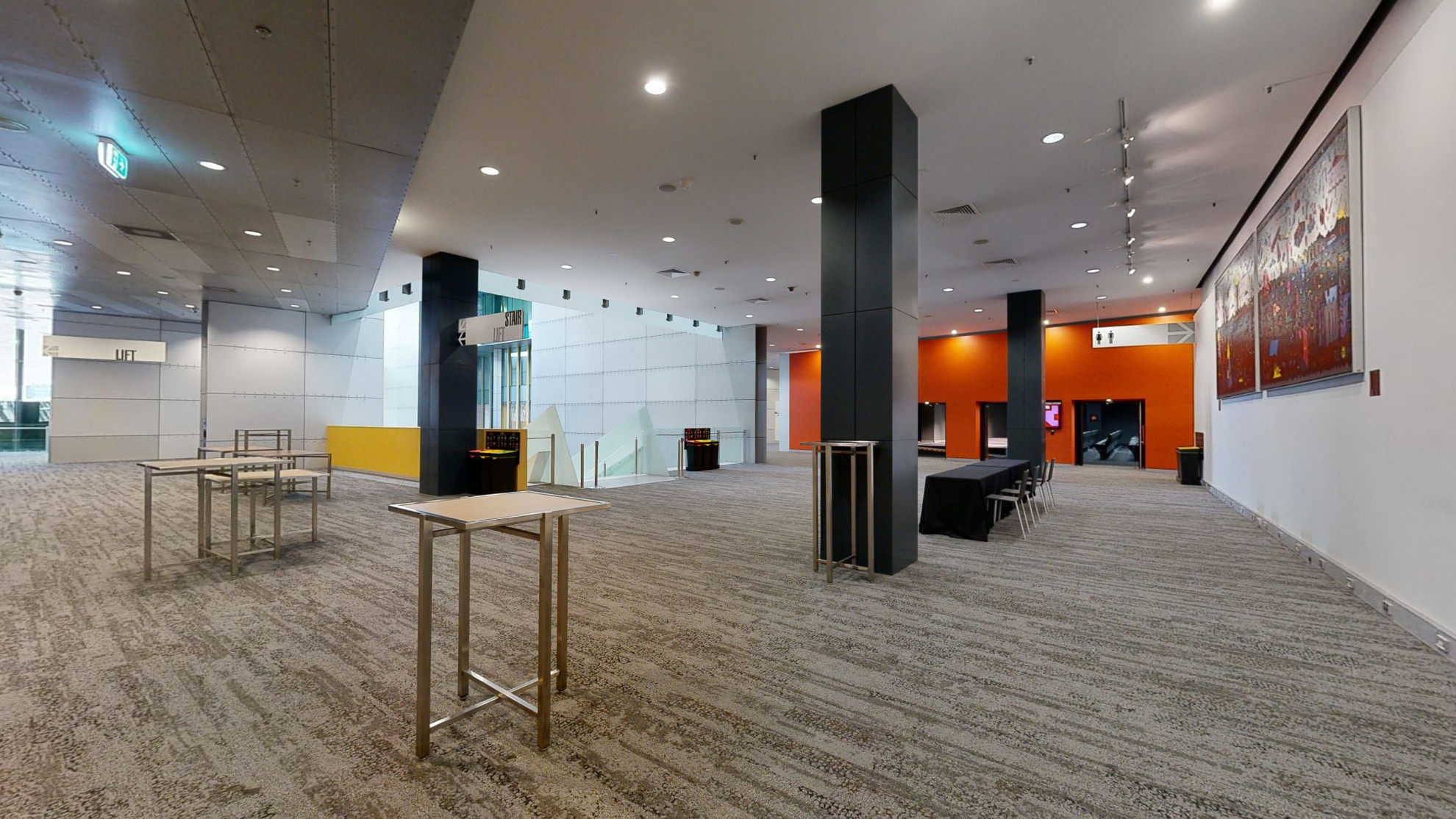 An empty foyer with orange walls, a prominent painting on the wall and scattered standing tables around with a main seated table and chairs facing the staircase coming up to the foyer.