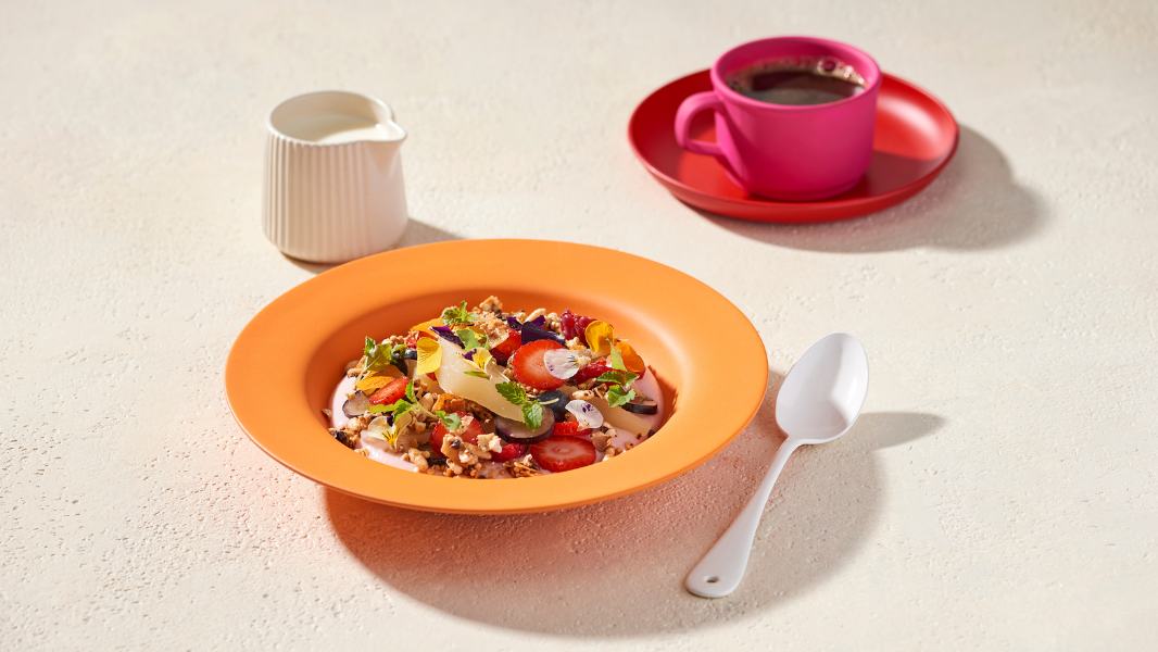 Strawberry gum yoghurt, poached pear and granola presented in a bright orange bowl with white spoon laying next to it. 