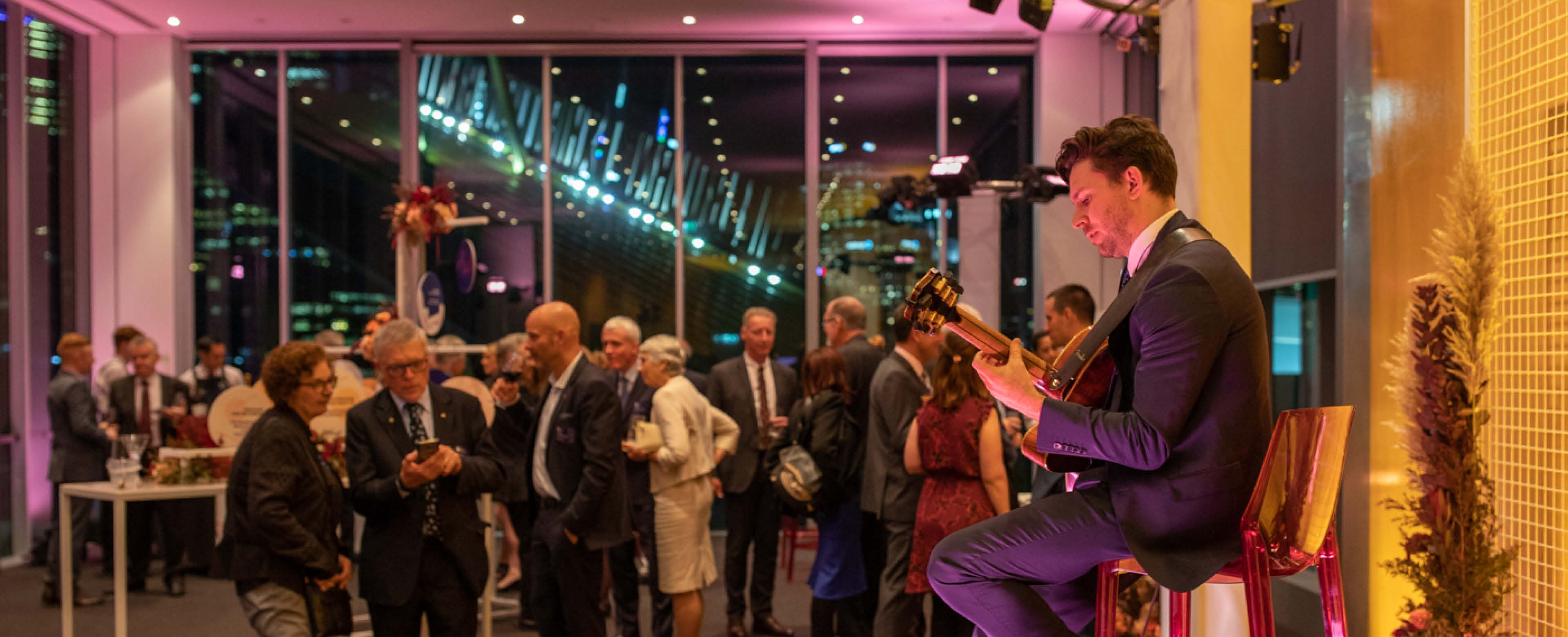 Musician playing guitar stands to the right of the image, performing to a crowd of people in formal attire socialising. 