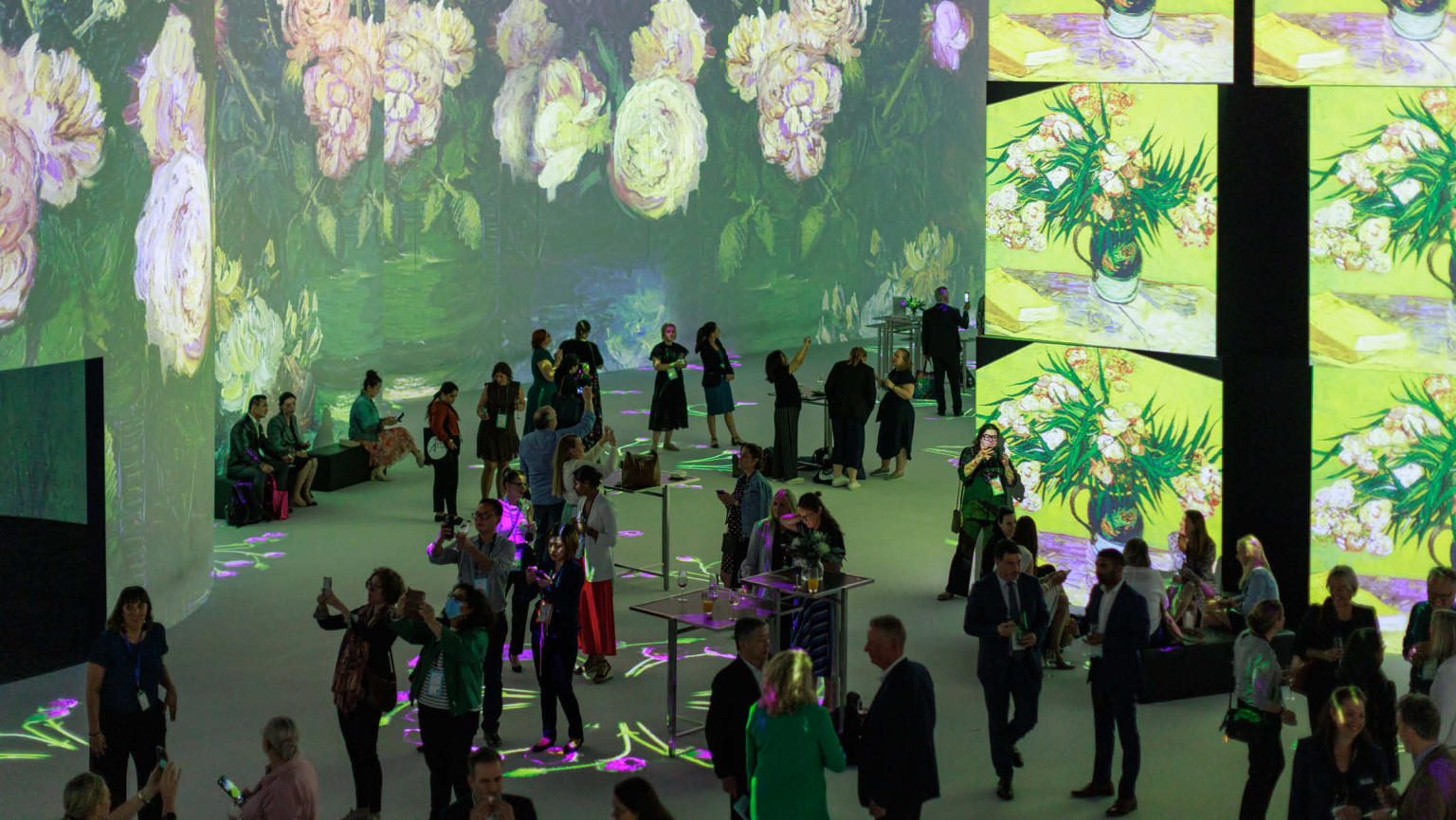 A large gathering of individuals congregated within a room adorned with expansive screens. The screens showcase vibrant yellow floral artwork, creating an immersive and visually captivating environment.
