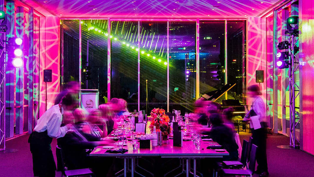 Long table with people sitting along either side in front of floor to ceiling windows. People sit at the table eating dinner with servers delivering them drinks. The room is filled with pink lighting. 