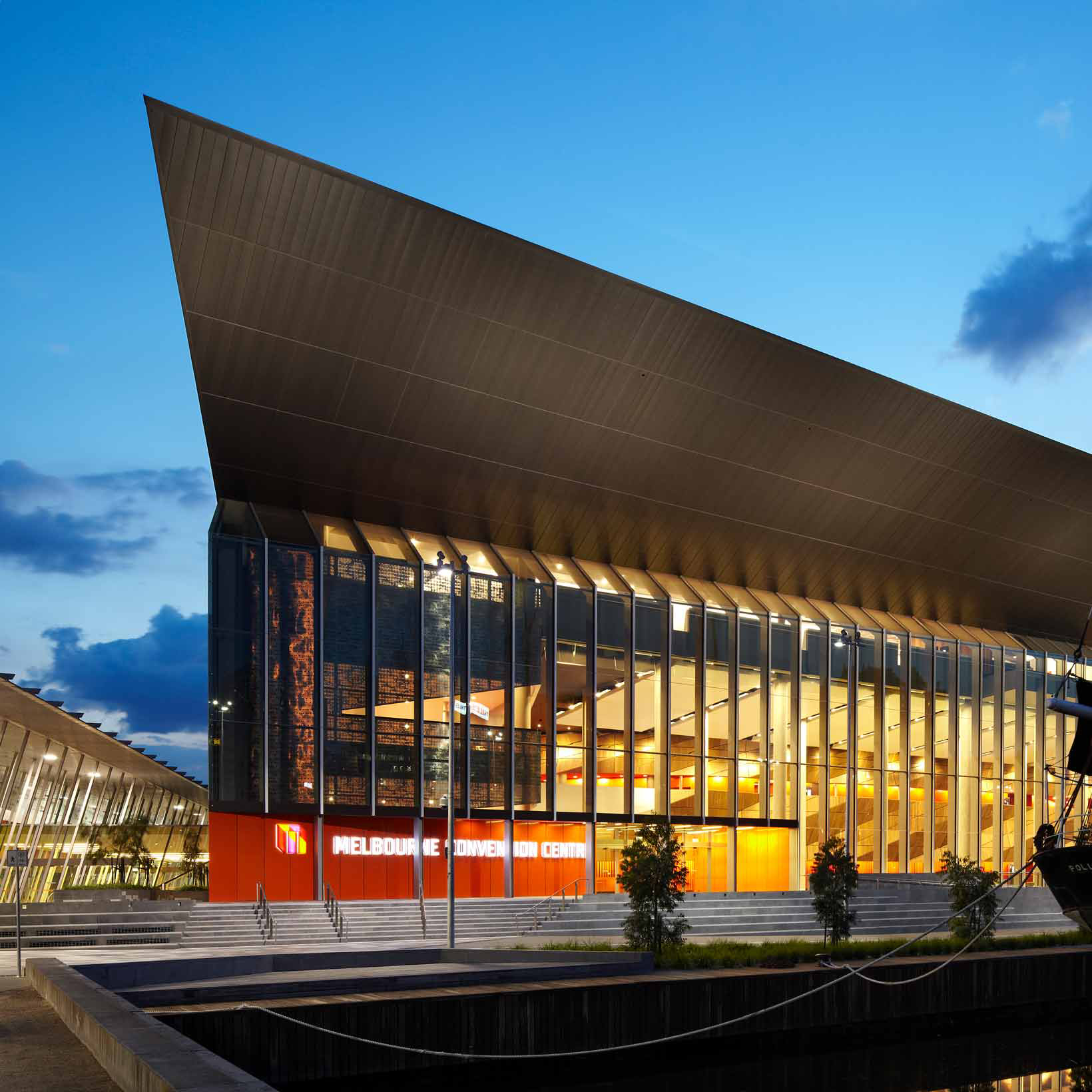 Deep blues and dark clouds fill the night sky with the Melbourne Convention Building lit up with yellow lights from inside in the foreground. 