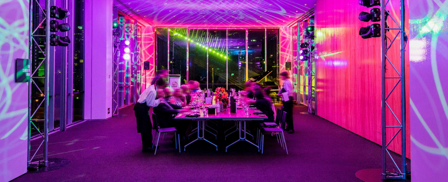 Long table with people sitting along either side in front of floor to ceiling windows. People sit at the table eating dinner with servers delivering them drinks. The room is filled with pink lighting. 