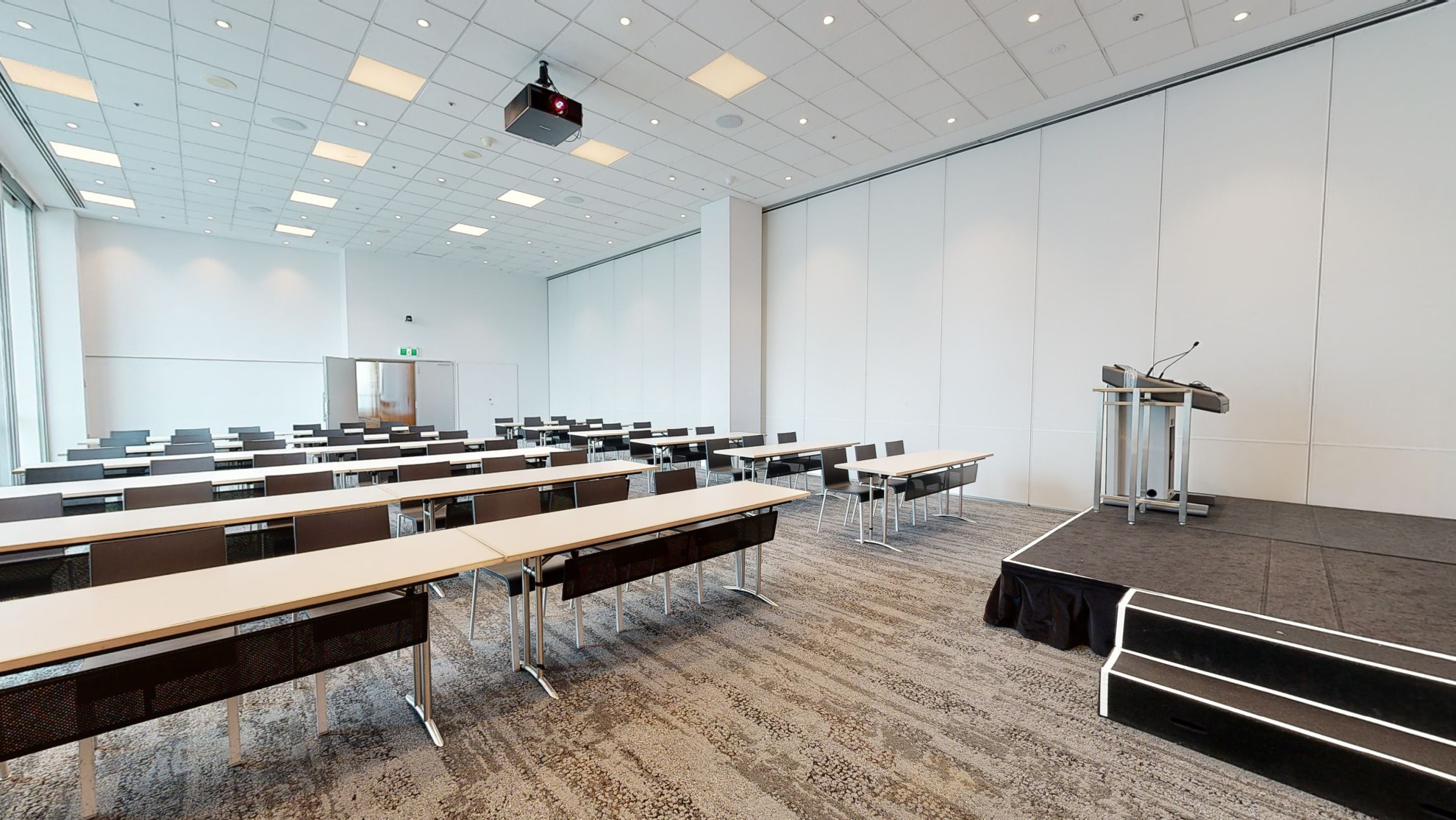 A professional conference or meeting room featuring a spacious layout, equipped with a stage and lectern and comfortable chairs for meetings and presentations. Table and chairs are set out in a classroom formation of rows. 